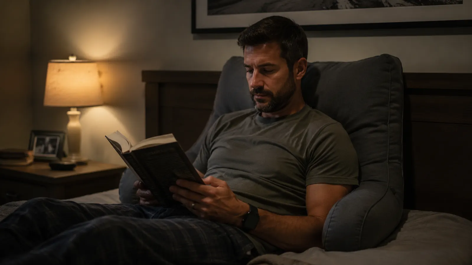 Man sitting up in bed against a grey armrest reading pillow, lamp glowing warm beside him, book open on his lap