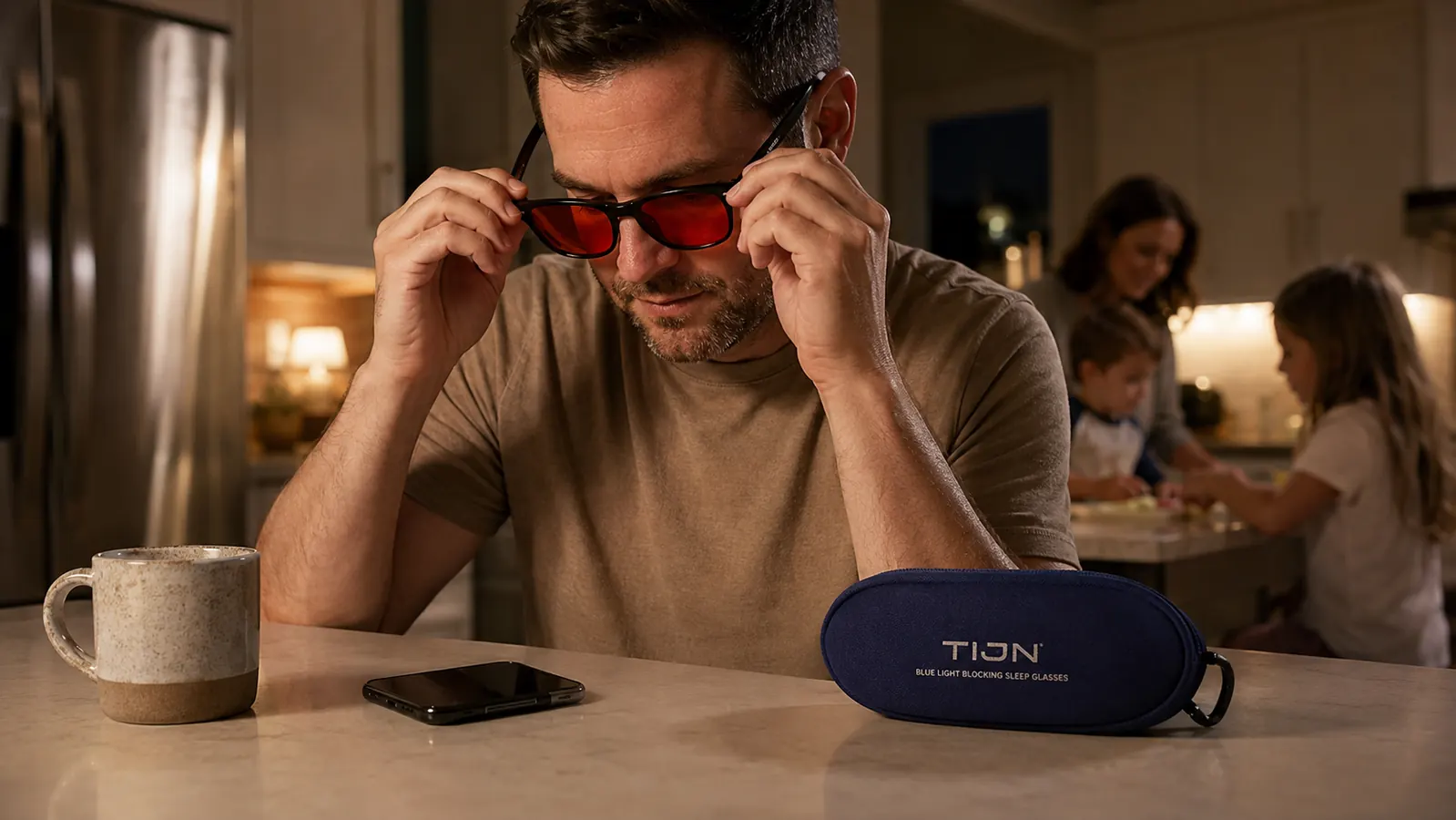 Close-up of hand putting on TIJN red-lens blue light blocking glasses at a kitchen counter with a phone nearby