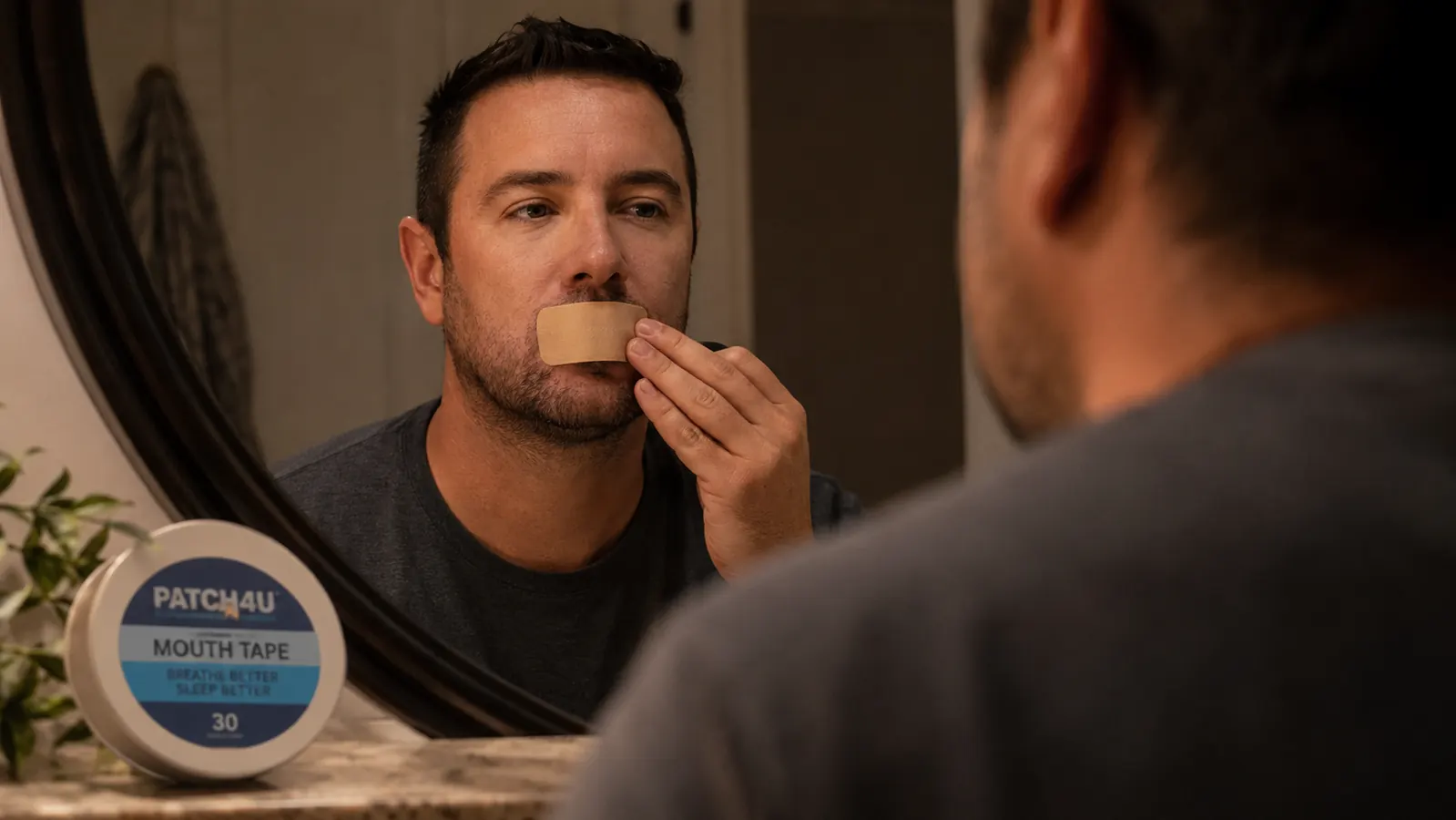 Hand applying a small vertical strip of mouth tape over closed lips in a bathroom mirror