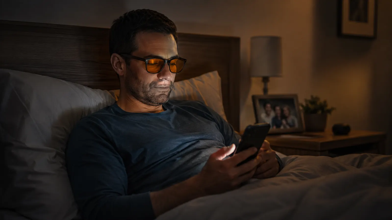 Man lying in bed at night wearing orange tinted glasses while looking at his phone with the lamp dimmed